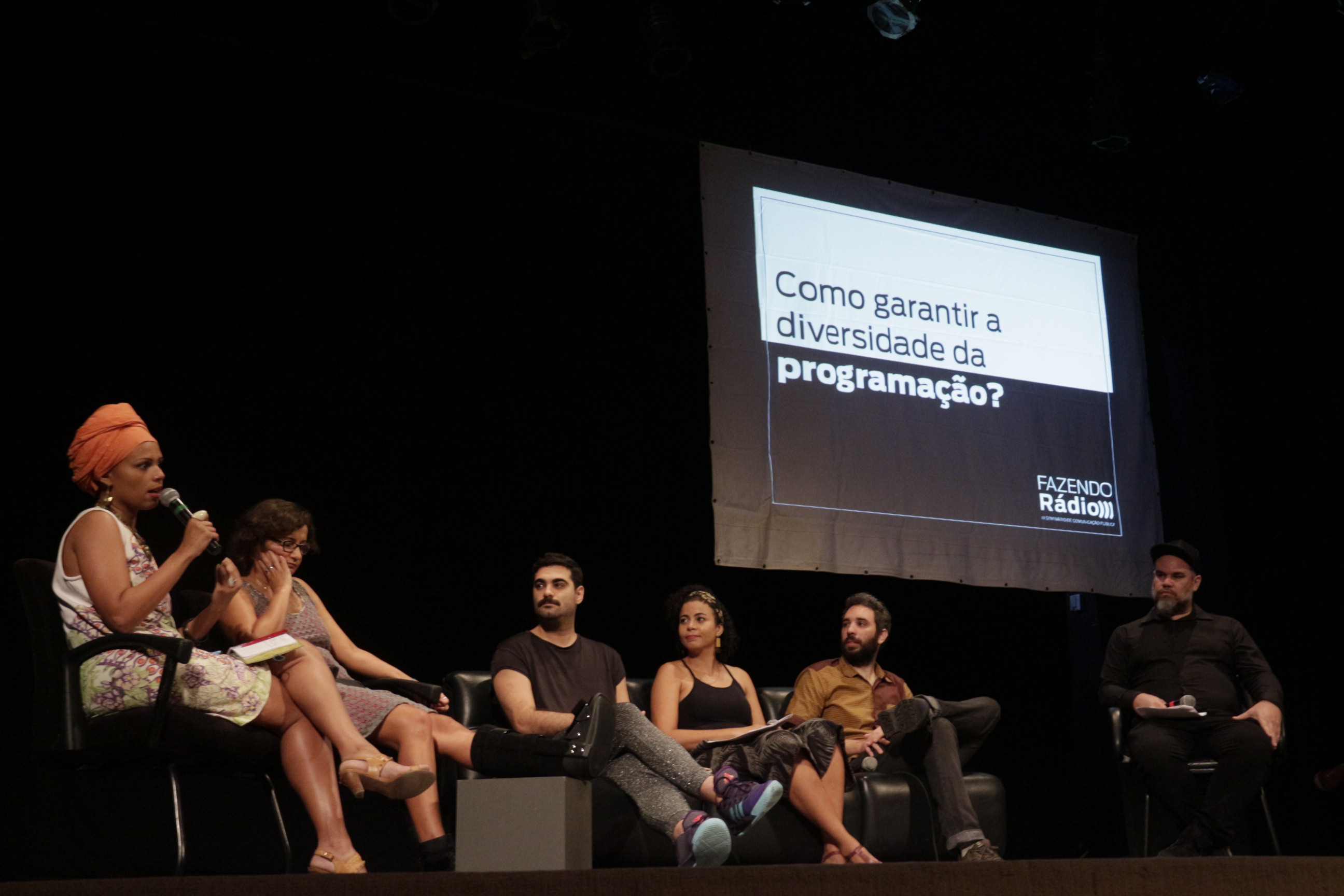 Palestrantes estão sentados em poltronas pretas no palco do teatro, da esquerda para a direita encontram-se: Juliana Nunes, Flavia Filipini, Paulo Proença, Nice Lima, Rodrigo Martins e Patrick Torquato, ao fundo lê-se um slide com a pergunta: como garantir a diversidade na programação?