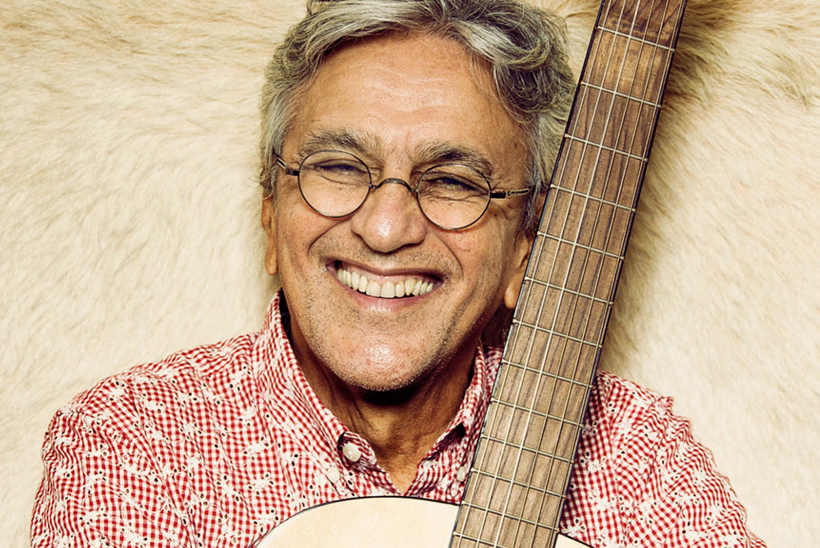 Caetano Veloso veste uma camisa quadriculada vermelho e sorri, abraçado a um violão. Foto: 