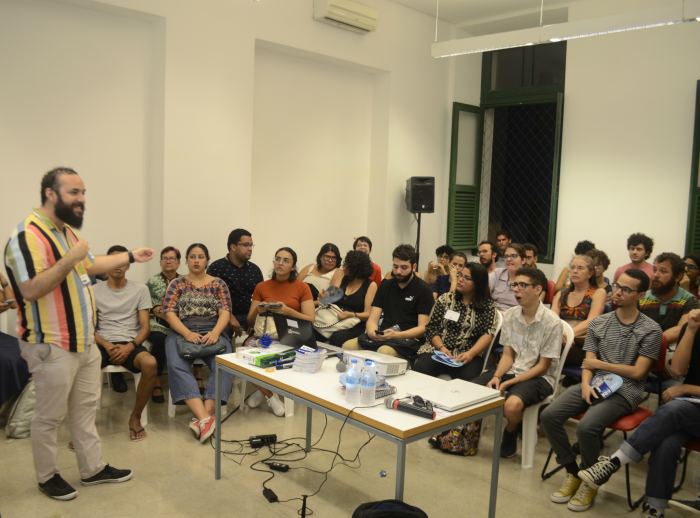 Em pé, no canto esquerdo, o oficineiro Tarcísio Camelo falando com os participantes da atividade, sentados a partir do centro, em direção ao canto direito.