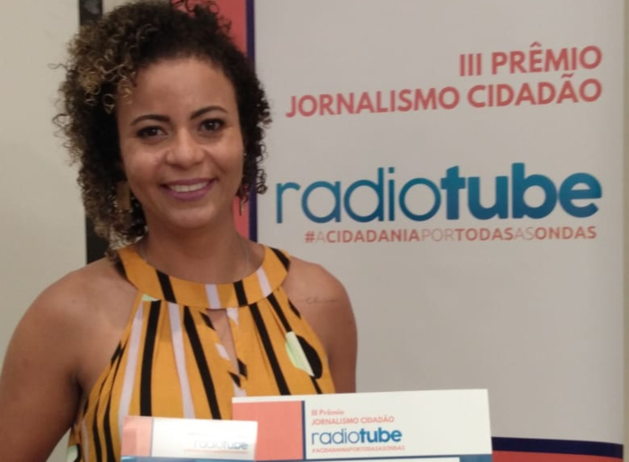 Imagem da radialista Nice Lima, sorrindo, segurando um prêmio e um certificado, olhando para nós.