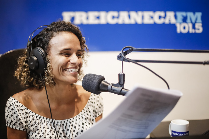 Mulher negra de cabelos cacheados em um estúdio de gravação. Ela está com fones de ouvido e tem um microfone  a sua frente. Seu semblante é feliz e ela sorri. 