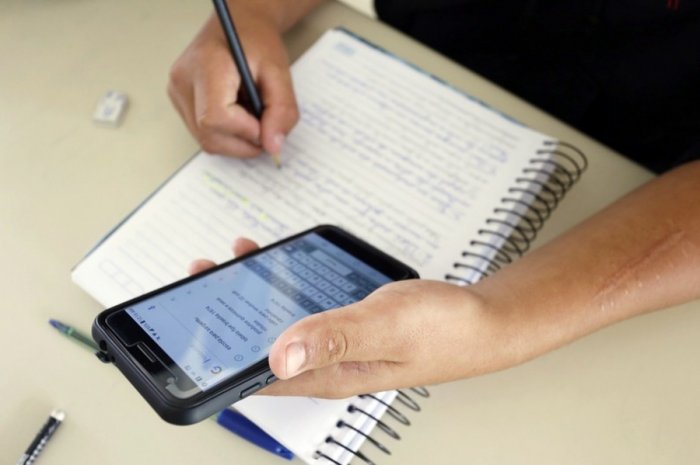 Na imagem, vê-se uma mão segurando um celular, a outra mão faz anotações em um caderno.
