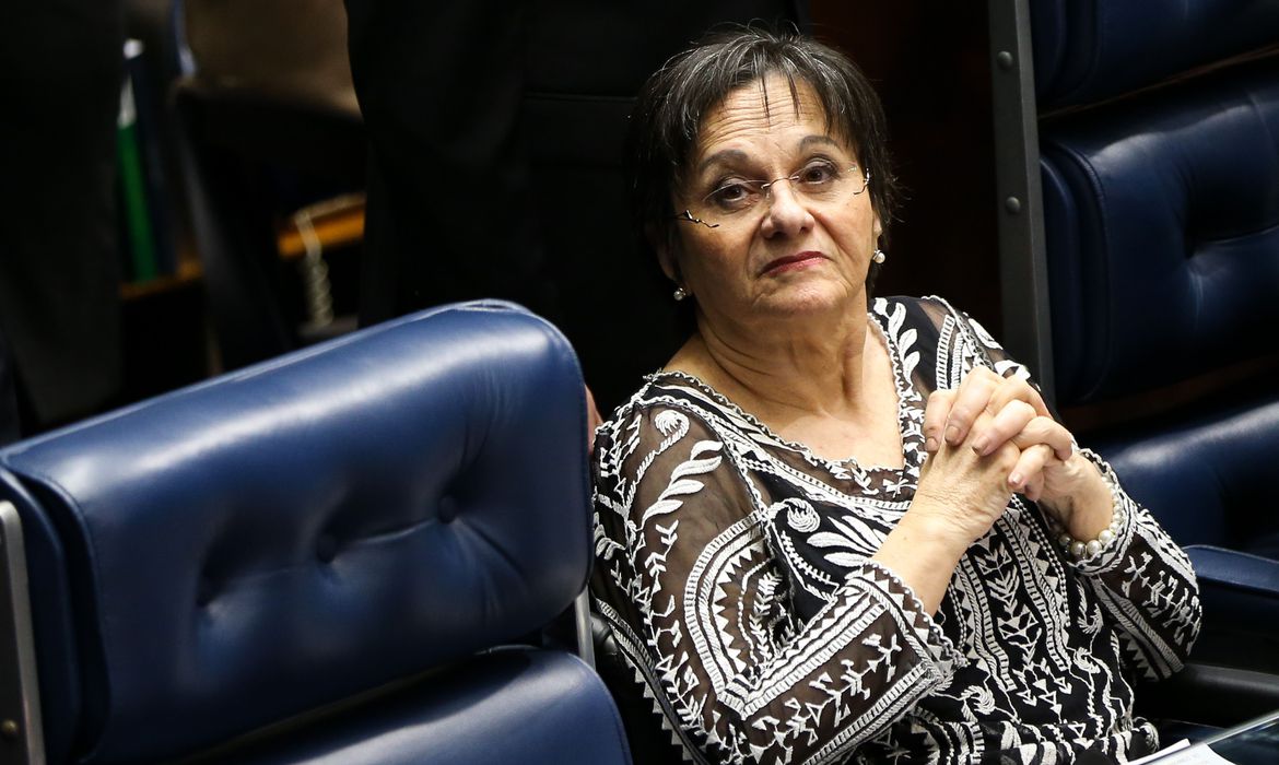 Foto de Maria da Penha, enquadramento até a cintura, mãos cruzadas, sentada ao lado de uma poltrona azul marinho. 