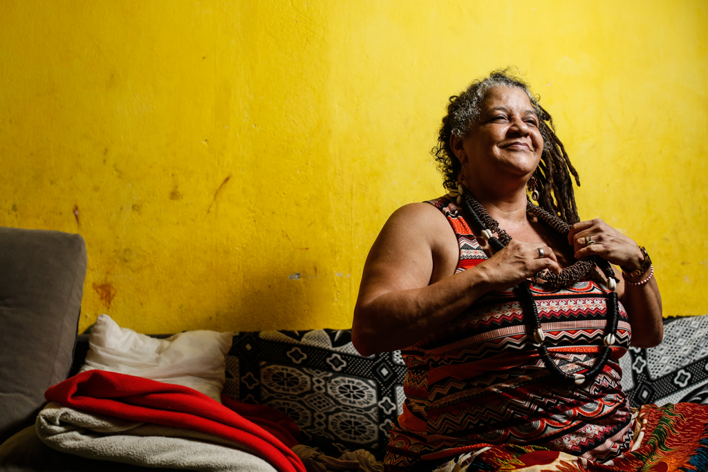 Foto de Mãe Beth de Oxum, mulher negra, com dreads, blusa estampada colorida, sentada em um sofá, com as duas mãos segurando um colar, olhando para o horizonte e esboçando um sorriso. Ao fundo, uma parede amarela.