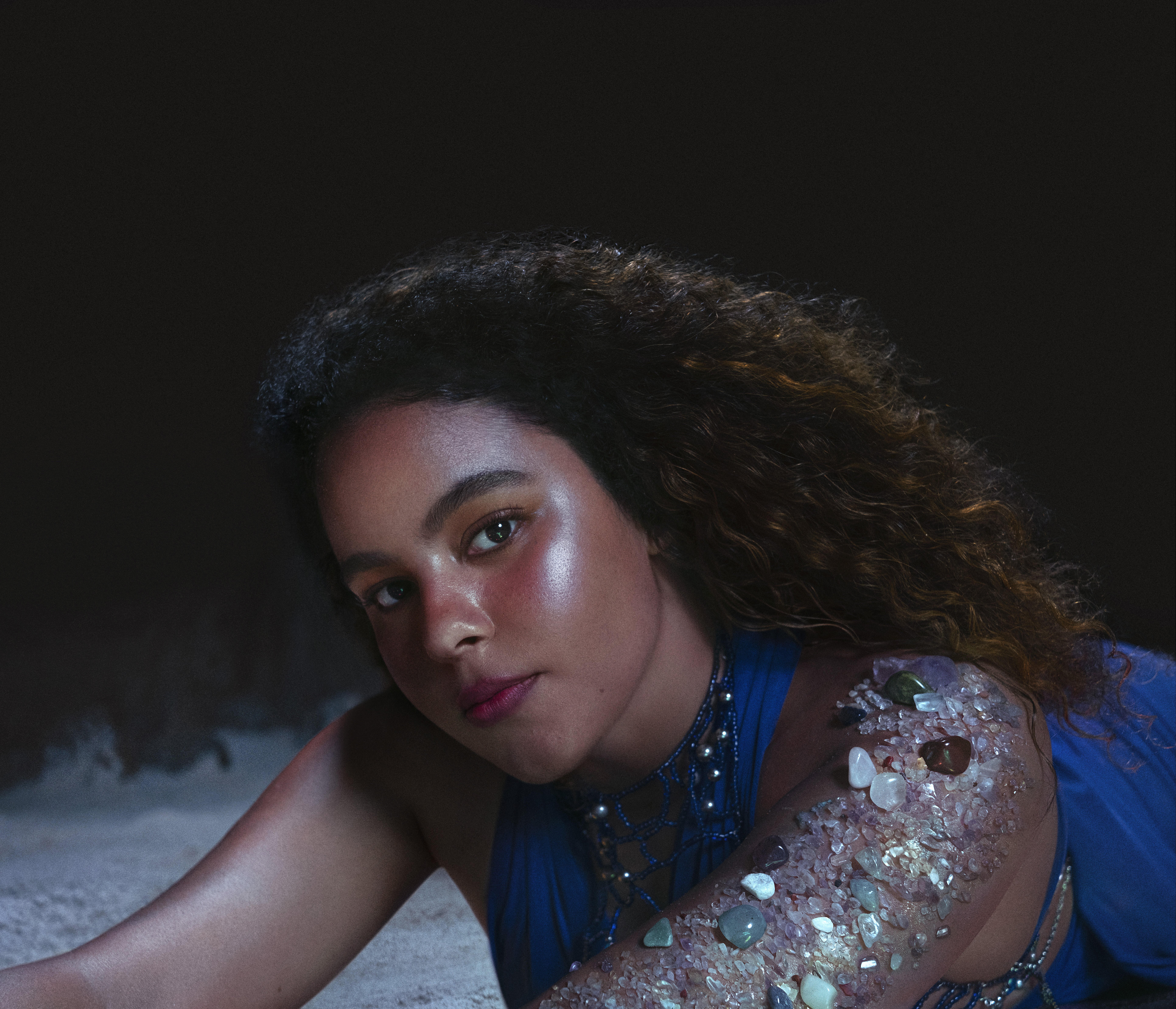 Mulher de pele negra clara e cabelos cacheados, com expressão serena, está deitada sobre uma superfície clara. Ela usa uma roupa azul com detalhes em miçangas e colares. Seu braço direito, em destaque na imagem, está coberto por cristais e pedras de diversas cores, como verde, branco e marrom, que brilham sob a luz. O fundo é escuro, o que contrasta com a iluminação suave que realça o brilho da pele e das pedras. A foto tem um clima artístico e delicado. Crédito da imagem: Érico Toscano.
