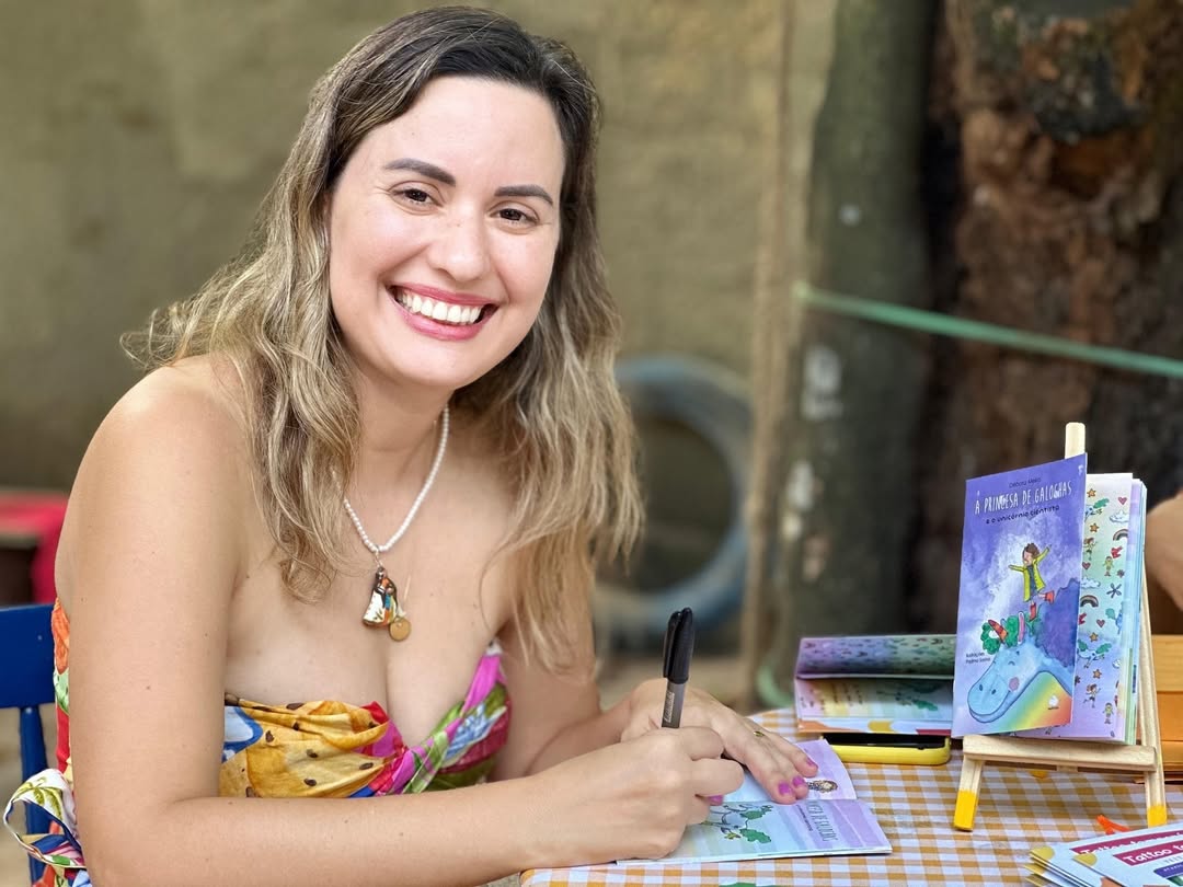 Mulher branca, de cabelos castanhos claros e ondulados, usa um vestido colorido de alças finas. Ela sorri amplamente para a câmera enquanto autografa um livro infantil. Está sentada à mesa coberta com uma toalha xadrez amarela e branca. Ao lado, está um cavalete com o livro A Princesa de Galochas, de capa roxa. Ao fundo, há árvores e um ambiente externo com clima acolhedor.