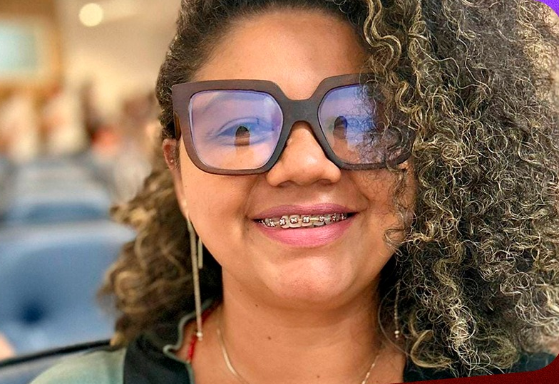 Mulher negra sorrindo em um ambiente interno, com poltronas azuis desfocadas ao fundo. Ela tem cabelos cacheados com luzes douradas, usa óculos grandes de armação escura e está com aparelho ortodôntico nos dentes. Veste uma blusa colorida e usa brincos longos e finos. A imagem está focada em seu rosto, transmitindo simpatia e confiança.