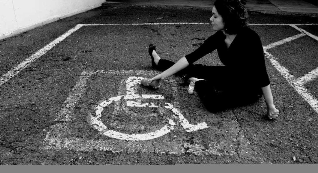 Imagem em preto e branco mostra uma mulher jovem sentada no chão de um estacionamento, dentro de uma vaga com o símbolo de acessibilidade pintado no asfalto. Ela está de pernas cruzadas, usa roupas escuras e segura uma lata de tinta spray, como se estivesse retocando o símbolo. A expressão no rosto é serena e reflexiva. A cena transmite uma mensagem simbólica sobre pertencimento e visibilidade.