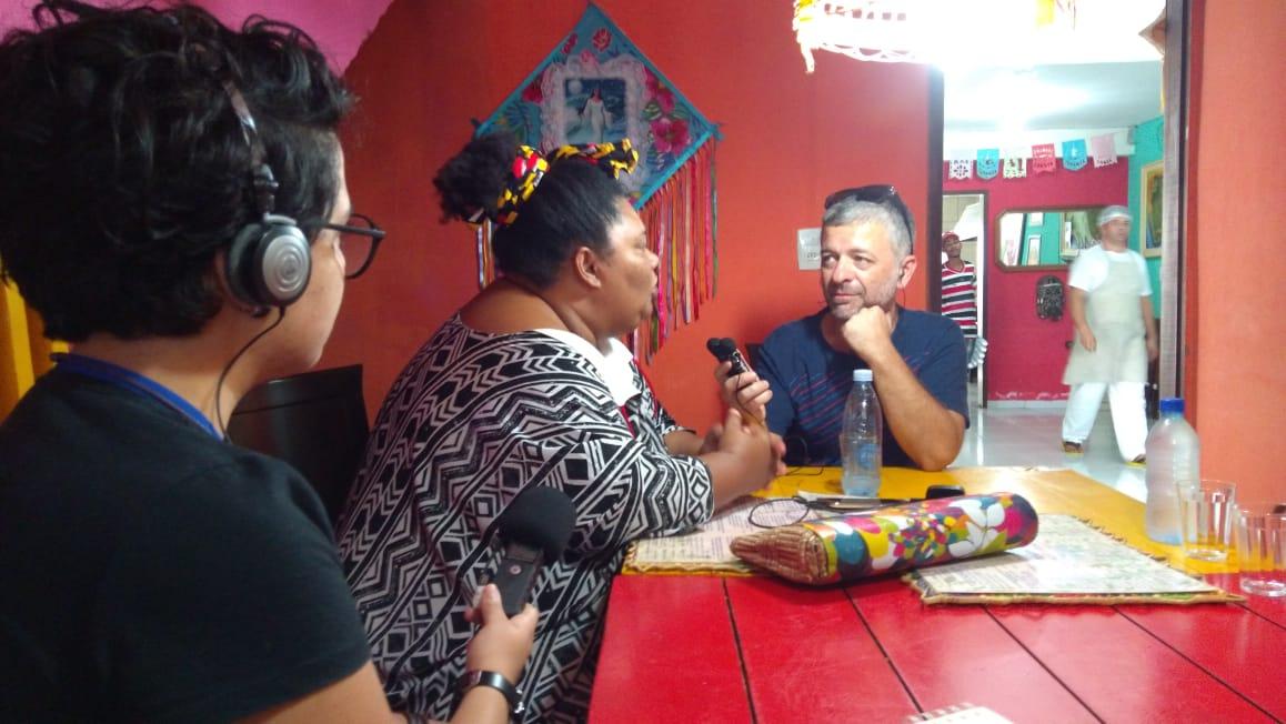 Kosta Kostov, Dona Carmem e Gabriele Alves estão sentados ao redor de uma mesa. Do lado esquerdo, Gabriele, usando fones de ouvido e um gravador de áudio, registra a entrevista. No centro, Dona Carmem olha para Kosta, que está à sua direita, enquanto responde a entrevista. Kosta observa Dona Carmem.