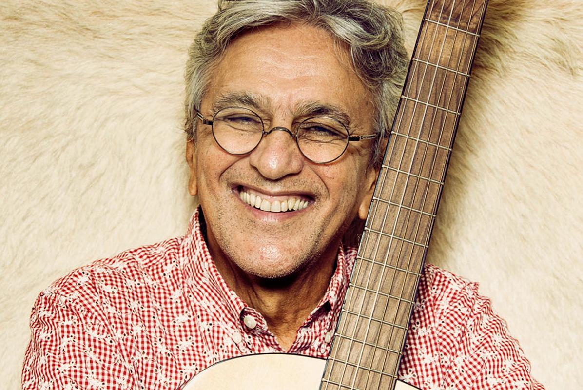 Caetano Veloso veste uma camisa quadriculada vermelho e sorri, abraçado a um violão. Foto: 