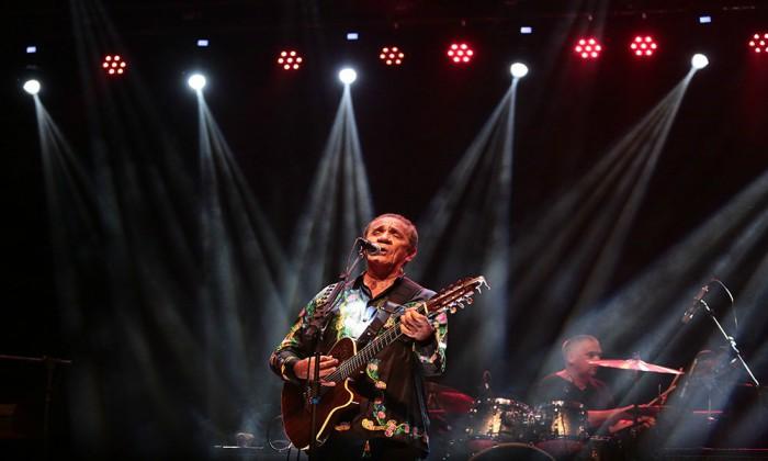 Vista frontal do cantor Zé Ramalho no centro do palco, empunhando o seu violão. Ao fundo, à direita, o baterista de sua banda.