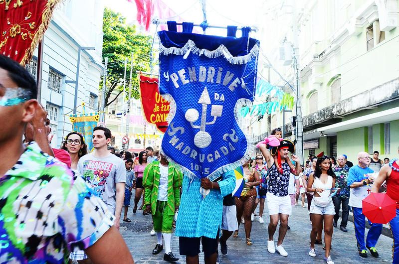 Na imagem, vê-se alguém segurando o estandarte do Pen Drive da Madrugada, durante seu desfile em 2019. Ao redor, algumas pessoas fantasiadas.