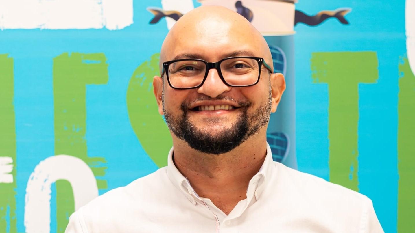 Foto de Bráulio Moura, careca, com barba, de óculos de grau preto, camisa social branca, em frente a um mural colorido.