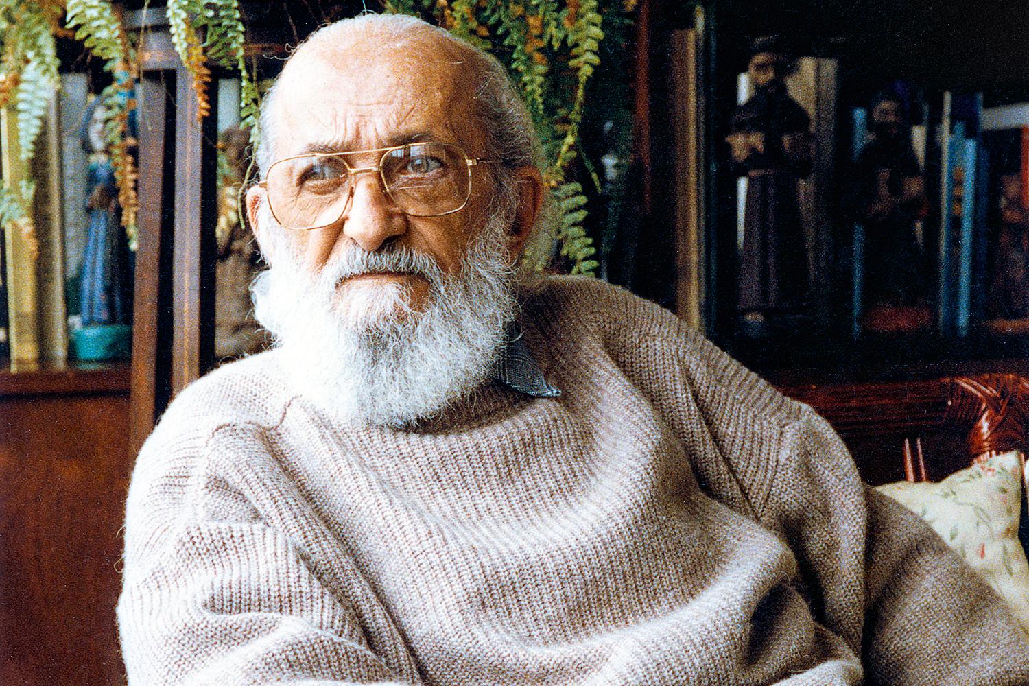 Foto de Paulo Freire, homem de barba branca, careca, de óculos, com um moletom bege. Ao fundo, uma estante cheia de livros.