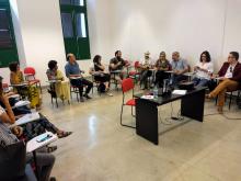 Representantes do Grupo de trabalho estão sentados, em reunião, em uma sala branca.