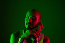 Na imagem, o cantor Lucas Torres está apoiado em uma das mãos, usando pulseiras e anéis, enquanto olha para a frente. Ele está iluminado por uma luz verde, em um fundo da mesma cor.