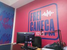 Foto mostra detalhe do estúdio da Frei Caneca FM, com parede em fundo rosa e letras azuis "Frei Caneca 101.5 FM"