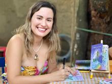 Mulher branca, de cabelos castanhos claros e ondulados, usa um vestido colorido de alças finas. Ela sorri amplamente para a câmera enquanto autografa um livro infantil. Está sentada à mesa coberta com uma toalha xadrez amarela e branca. Ao lado, está um cavalete com o livro A Princesa de Galochas, de capa roxa. Ao fundo, há árvores e um ambiente externo com clima acolhedor.