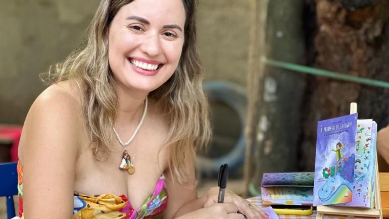 Mulher branca, de cabelos castanhos claros e ondulados, usa um vestido colorido de alças finas. Ela sorri amplamente para a câmera enquanto autografa um livro infantil. Está sentada à mesa coberta com uma toalha xadrez amarela e branca. Ao lado, está um cavalete com o livro A Princesa de Galochas, de capa roxa. Ao fundo, há árvores e um ambiente externo com clima acolhedor.