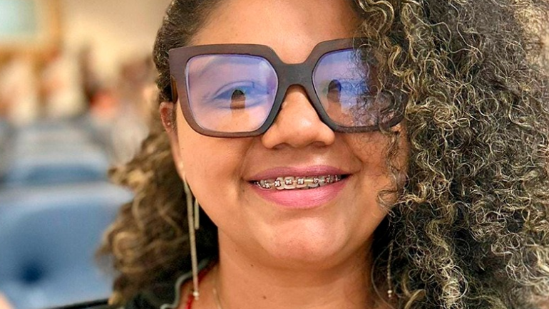 Mulher negra sorrindo em um ambiente interno, com poltronas azuis desfocadas ao fundo. Ela tem cabelos cacheados com luzes douradas, usa óculos grandes de armação escura e está com aparelho ortodôntico nos dentes. Veste uma blusa colorida e usa brincos longos e finos. A imagem está focada em seu rosto, transmitindo simpatia e confiança.